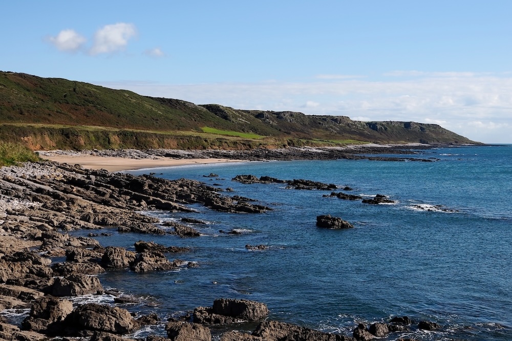 Photograph of a bay showing a rocky shoreline and sandy beach behind it.