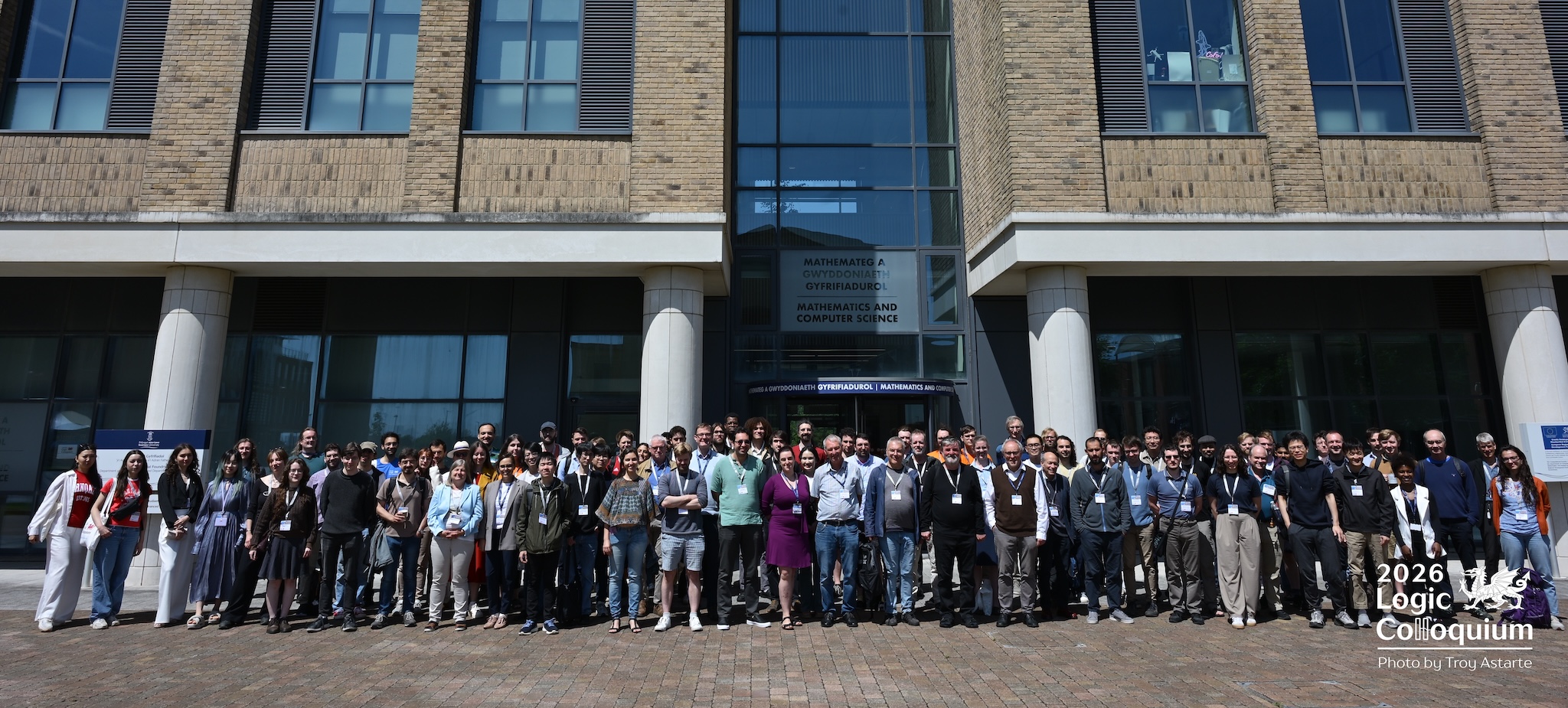 photograph shows a large number of people standing in a few rows arrayed outside an academic building. The sun is shining.