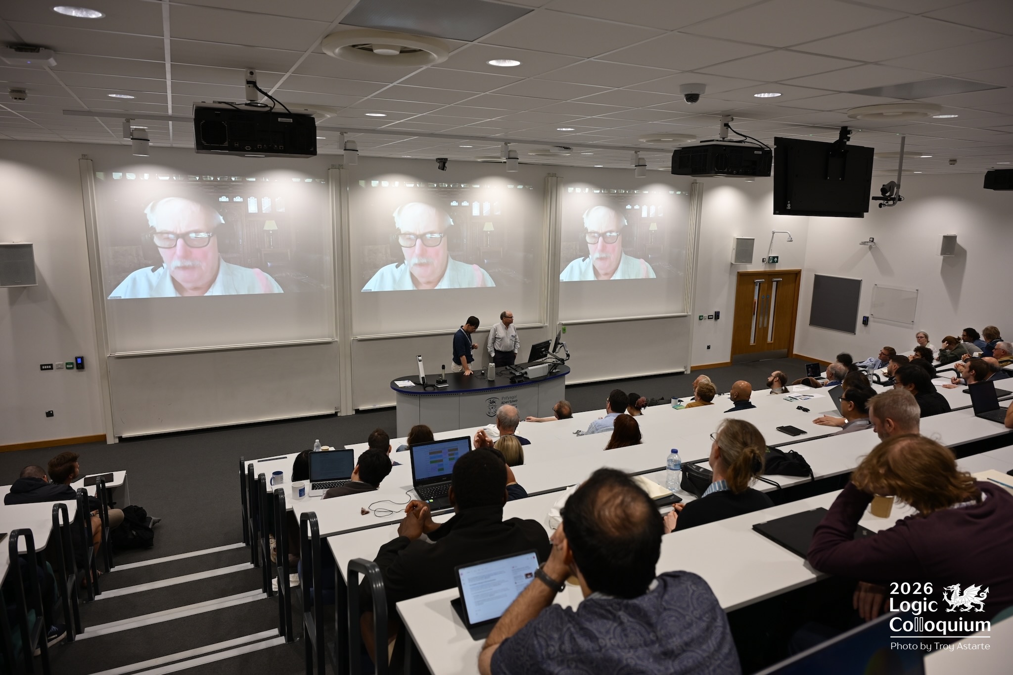 photograph interior of lecture theatre from the back row. Attendants watch three large screens showing the face of John Burgess, mid-lecture.