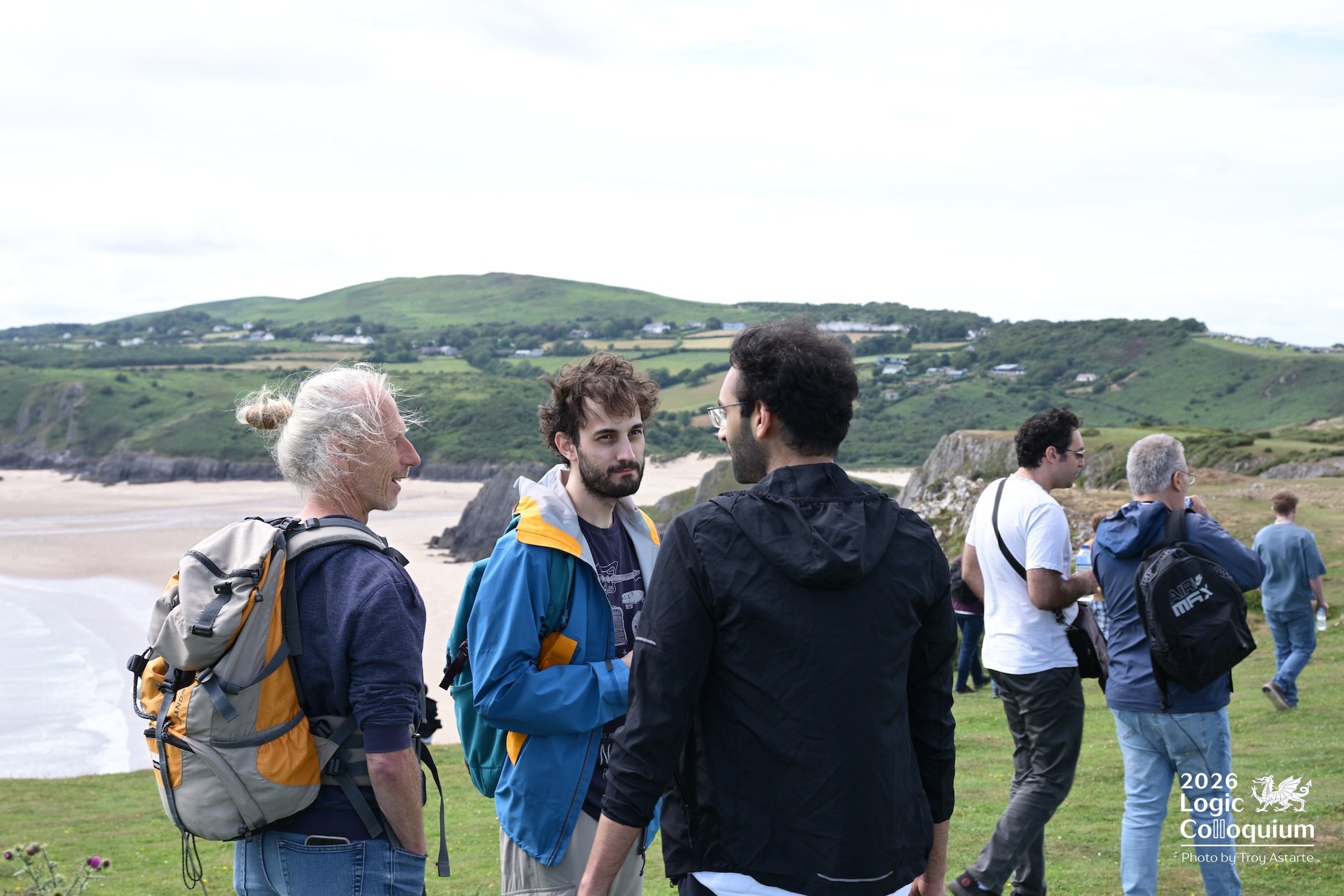 Photograph of three serious academics in deep conversation while the winds of the Gower blow their hair, and others in the background begin to drift towards the paths leading down to the Bay.